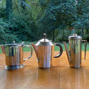 May include: Three polished silver-toned metal vessels arranged in a row. The left is a short, wide teapot with a curved handle. The centre is a medium-height teapot with a dark handle and lid. The right is a tall, slender coffee pot with a flat lid.