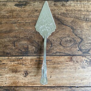 May include: Silver pie server with a decorative floral pattern on the blade and handle. The server is made of metal and has a long, slender handle. The server is resting on a wooden surface.