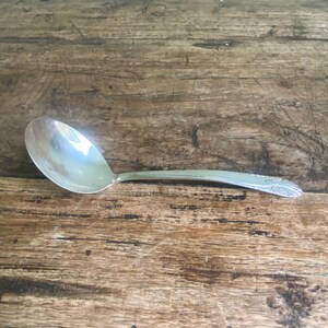 May include: A silver spoon with a rounded bowl and a long, slender handle. The handle features a decorative floral pattern near the end. The spoon is on a weathered wooden surface.