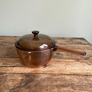 May include: A vintage brown glass saucepan with a matching lid and a long handle. The cookware is made of transparent glass, allowing visibility of the contents. The saucepan is sitting on a wooden surface.