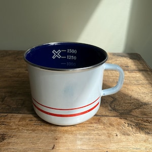 May include: White enamel mug with a blue interior and a red stripe. The mug has a light blue handle and measurement markings for 1000, 1250, and 1500 ml. The mug is on a wooden surface.