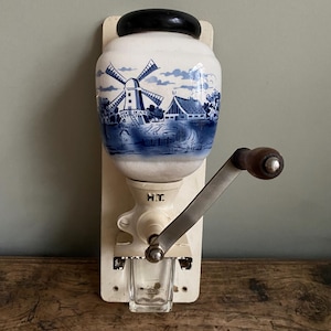 May include: Vintage white and blue ceramic coffee grinder with a black top and wooden handle. The ceramic container has a blue windmill and landscape design. A small glass cup is below the grinding mechanism.