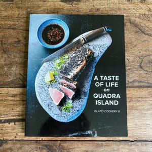 Peut inclure: Un livre de cuisine intitulé "A Taste of Life on Quadra Island" avec une couverture sombre. La couverture présente une assiette de nourriture avec un couteau, un petit bol bleu de sauce et du poisson tranché. Le livre fait partie de la série Island Cookery III.