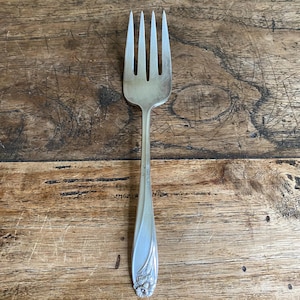 May include: A silver fork with four prongs, set against a wooden background. The handle has a floral pattern at the base. The fork is a vintage item, showing signs of use.