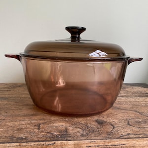 May include: A brown glass casserole dish with a matching lid. The dish has two side handles and a dark knob on the lid. The glass is transparent, allowing a view of the inside. The dish is sitting on a wooden surface.