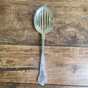 May include: A silver slotted spoon with an oval head and a long, ornate handle. The spoon's head has several vertical slots. The handle features decorative floral patterns. The spoon is made of metal and is likely used for serving.