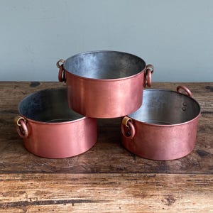 May include: Three copper-coloured metal bowls with rounded sides and small handles. The bowls are stacked on a wooden surface, with the top bowl centered above the other two. The interior of the bowls is a lighter silver colour.