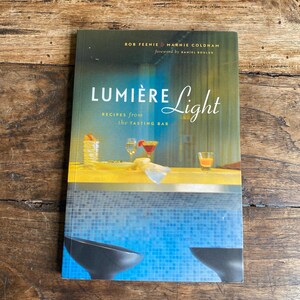 May include: A cookbook titled "Lumière Light" with the subtitle "Recipes from the Tasting Bar." The cover displays cocktails and glassware. The title is in a stylish font, with the authors' names at the top. The book is on a wooden surface.