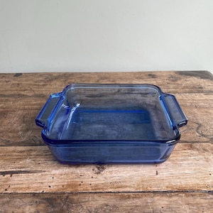 May include: A rectangular, sapphire blue glass baking dish with two handles. The dish is empty and sits on a wooden surface. The glass is transparent. A kitchenware item.