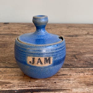 Peut inclure: Pot à confiture en céramique bleue avec couvercle et petite poignée. Le mot "JAM" est imprimé sur le devant. Le pot est posé sur une surface en bois.