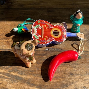 May include: A collection of four ornaments including a red guitar with floral patterns and the text Feliz Navidad, a green and blue parrot, a small tan animal figurine with brown markings, and a bright red chilli pepper hanging ornament.