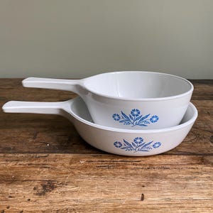 May include: Two white handled bowls, one stacked inside the other. Each bowl has a blue floral pattern. The bowls are on a wooden surface.