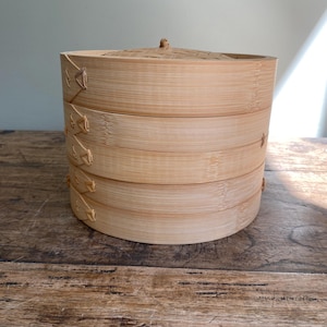 May include: A natural bamboo steamer with a lid, featuring four stacked tiers. The light brown, woven bamboo construction is held together with woven accents. The steamer sits on a wooden surface, suggesting a kitchen or dining setting.