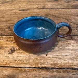 May include: A handcrafted ceramic mug. The exterior is brown, while the interior is a striking blue. The mug has a rounded shape, a comfortable handle, and a glossy finish. Ideal for tea or coffee, it has a rustic design.