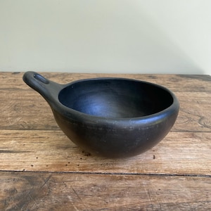 May include: A black ceramic bowl with a handle. The bowl is round with a wide opening and a smooth, matte finish. The handle has a small hole at the end. The bowl is sitting on a wooden surface.