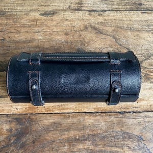 May include: A black, cylindrical leather case with a handle and two straps. The case has a textured surface and brown stitching. The straps are secured with silver-coloured metal studs. The case is on a wooden surface.