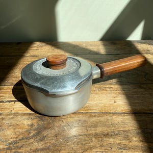 May include: A small, vintage aluminium saucepan with a wooden handle and lid. The lid has a wooden knob. The saucepan is on a wooden surface, with sunlight creating shadows across the scene.