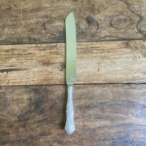 Peut inclure: Un couteau à gâteau argenté avec un bord dentelé et une poignée ornée. La lame est lisse et réfléchissante, tandis que la poignée présente un motif floral détaillé. Le couteau est présenté sur une surface en bois rustique.