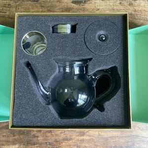 May include: A black glass teapot set, including a lid, infuser, and a container with the text "FORTNUM", presented in a box. The teapot features a curved spout and handle. The set is set in a dark grey foam insert within a gold-edged box.