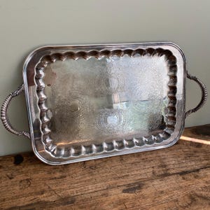 Peut inclure: Un plateau de service rectangulaire en argent avec des motifs floraux ornés et des bords festonnés. Le plateau a deux poignées incurvées et repose sur une surface en bois. Le plateau est probablement vintage ou antique.