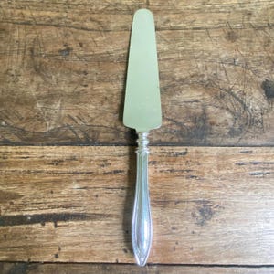 May include: A silver cake server with a broad, flat blade and a decorative handle. The blade is a pale green colour, and the handle has an ornate design. The server is made of metal and is likely vintage.