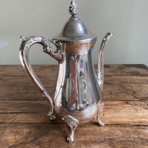 May include: A silver-plated coffee pot with a decorative handle and spout. The pot has a domed lid with a finial and rests on three ornate feet. The polished surface reflects the surrounding environment, creating a vintage aesthetic.