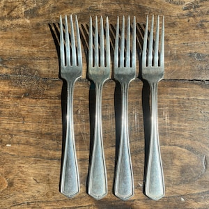 May include: Four silver forks with long, slender tines and decorative handles are arranged vertically on a wooden surface. The forks have a classic design, reflecting light and casting shadows. The wooden surface has a warm, natural tone.