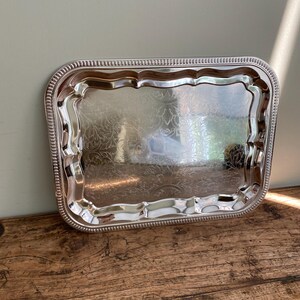 May include: A rectangular silver serving tray with a decorative floral pattern etched into the surface. The tray has a scalloped edge and a beaded border. The tray is sitting on a wooden surface.