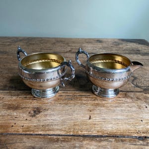 May include: Two antique silver-plated serving pieces. One is a sugar bowl with two handles, and the other is a creamer with a spout. Both have a gold-coloured interior and are set on a wooden surface.