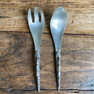 Peut inclure: Une paire de couverts à salade argentés vintage. L'un est une fourchette à trois dents, l'autre une cuillère. Les deux ont des manches finement sculptés avec un motif tribal. Les couverts sont présentés sur une surface en bois.