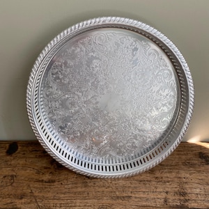 Peut inclure: Un plateau de service rond argenté avec un motif floral orné au centre. Le plateau a un bord décoratif en forme de corde et une bordure ajourée. Le plateau en métal est adapté pour servir des aliments et des boissons.