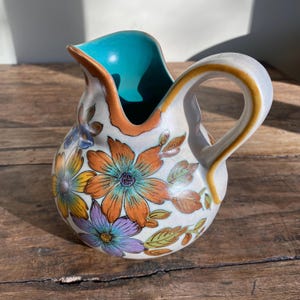 May include: A white ceramic jug with a turquoise blue interior and a floral design. The flowers are orange, yellow, purple, and blue. The jug has a brown handle.