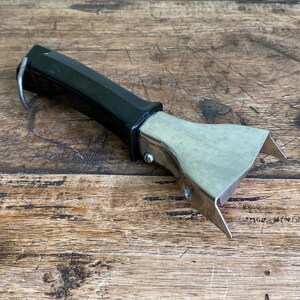 Peut inclure: Un lève-pot vintage à manche noir avec une tête en acier inoxydable. Le lève-pot est doté d'une boucle pour l'accrocher et est conçu pour saisir le bord d'une casserole chaude. L'outil est présenté sur une surface en bois.