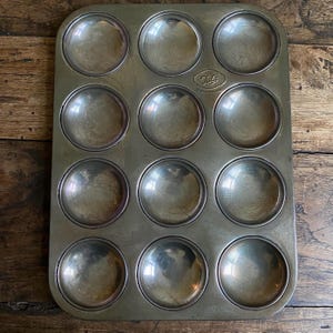May include: A vintage muffin tin with twelve circular molds. The tin is made of a tarnished, silver-coloured metal. The word "Tala" is stamped on the top of the tin. The tin is on a wooden surface.