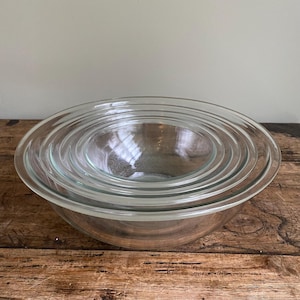 Peut inclure: Un ensemble de saladiers en verre transparent de différentes tailles, emboîtés les uns dans les autres. Les bols sont ronds et présentent un design simple et classique. L'image est prise sur une surface en bois.