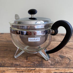May include: A clear glass Bodum tea infuser with a stainless steel lid and filter. The tea infuser has a black handle and a black knob on the lid. The Bodum logo is visible on the front of the infuser. The infuser sits on a wooden surface.