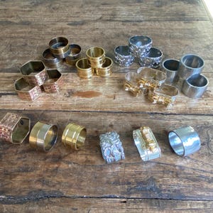 May include: A collection of assorted metal napkin rings in various finishes including gold, silver, and copper tones. The set features smooth bands, textured woven patterns, ornate embossed designs, and decorative bow accents in different shapes.