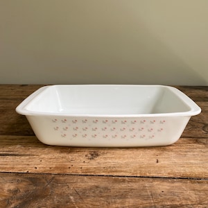May include: Rectangular white baking dish with a floral pattern of small pink and grey flowers. The dish has a slightly flared rim and is set on a wooden surface. A vintage kitchenware item.