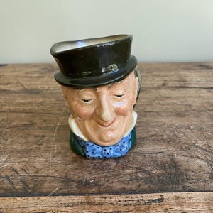 May include: A ceramic character jug shaped like a person wearing a black top hat, a white collared shirt, and a blue bowtie with black polka dots. The figure has a light skin tone, closed eyes, and a subtle smile, with a dark green coat collar visible.