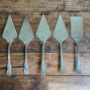 May include: Five silver-toned metal cake servers arranged in a row. Four servers feature triangular blades with intricate embossed floral patterns and ornate handles, while the fifth server has a rectangular blade with a similar detailed design.