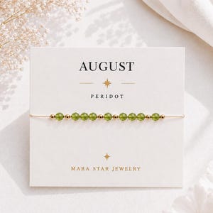 May include: A delicate bracelet featuring small, faceted green peridot beads and tiny gold-toned metal spacers, displayed on a white card printed with the text AUGUST, PERIDOT, and MARA STAR JEWELRY, accented with small star symbols.