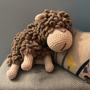 May include: A handmade, plush sheep toy with a white crocheted head and legs, and a brown, textured body. The sheep is resting on a blue patterned cushion, creating a cozy and inviting scene. The toy is designed to be soft and cuddly.