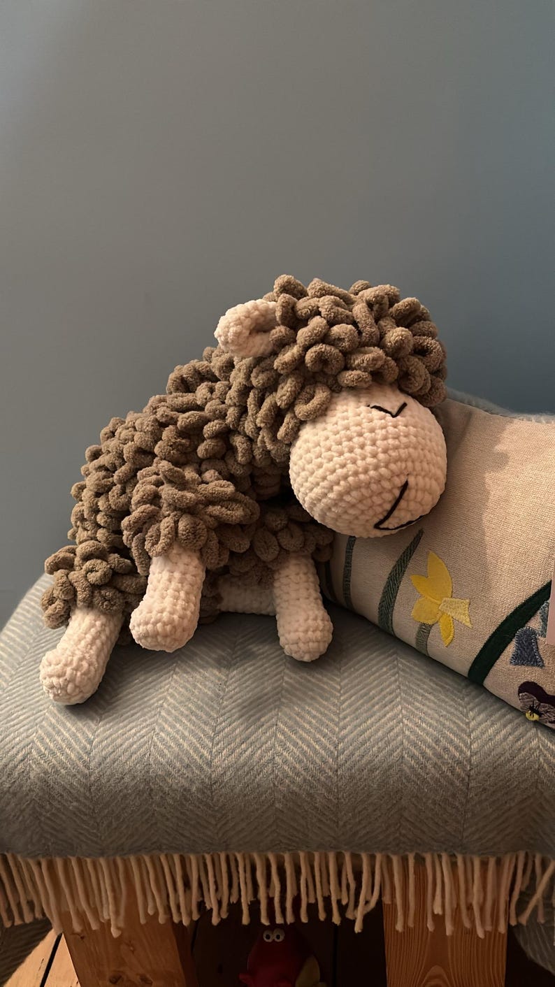 May include: A handmade, plush sheep toy with a white crocheted head and legs, and a brown, textured body. The sheep is resting on a blue patterned cushion, creating a cozy and inviting scene. The toy is designed to be soft and cuddly.
