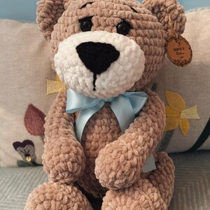 May include: A handmade, plush teddy bear in a soft brown color. The bear has a white muzzle, black eyes, and a light blue ribbon tied around its neck. A small tag is attached to the bear's ear. The bear is sitting on a patterned pillow.