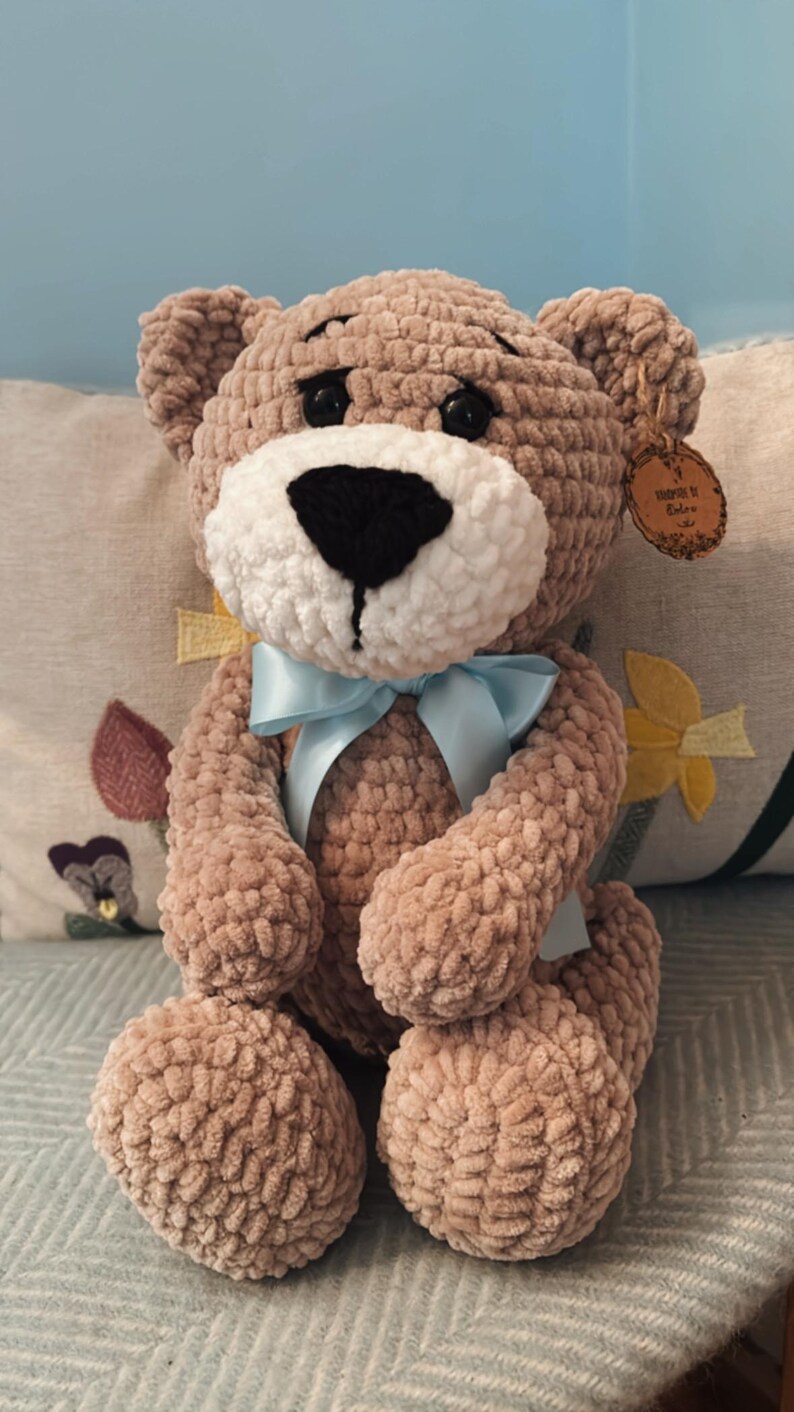 May include: A handmade, plush teddy bear in a soft brown color. The bear has a white muzzle, black eyes, and a light blue ribbon tied around its neck. A small tag is attached to the bear's ear. The bear is sitting on a patterned pillow.