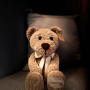 May include: A handmade, light brown teddy bear with a white ribbon tied around its neck. The bear has dark brown eyes and a dark brown nose. The word "Summer" is embroidered on one of its paws. The bear is sitting on a gray surface.