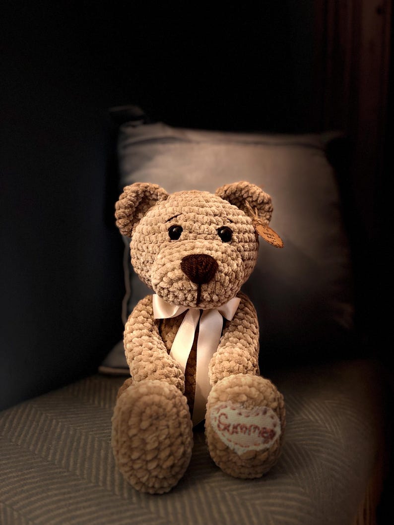 May include: A handmade, light brown teddy bear with a white ribbon tied around its neck. The bear has dark brown eyes and a dark brown nose. The word "Summer" is embroidered on one of its paws. The bear is sitting on a gray surface.
