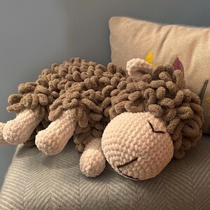 May include: A handmade, plush sheep toy in shades of brown and white. The sheep is lying down, with a fluffy, textured body and a smiling face. The toy is made of soft, chunky yarn, perfect for cuddling.