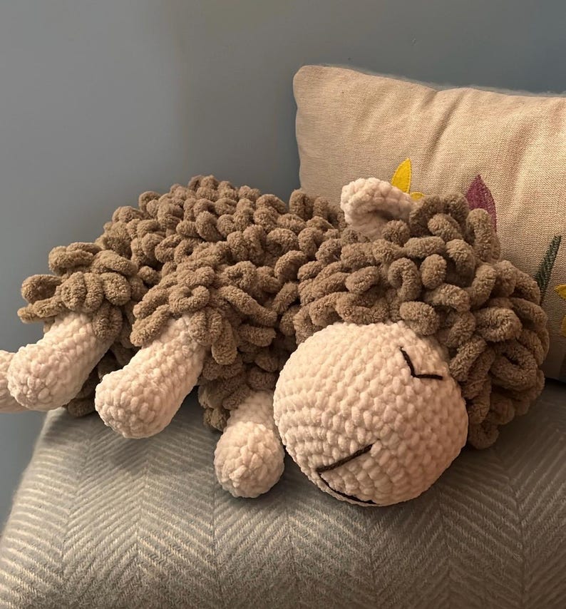 May include: A handmade, plush sheep toy in shades of brown and white. The sheep is lying down, with a fluffy, textured body and a smiling face. The toy is made of soft, chunky yarn, perfect for cuddling.