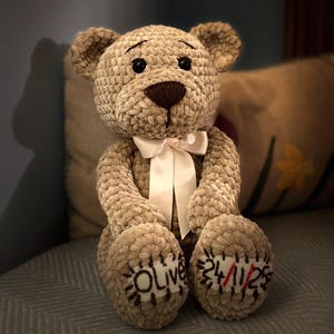May include: A handmade, plush teddy bear in a soft, textured beige fabric. The bear has a brown snout and black eyes, with a white ribbon tied around its neck. The bear's paws are embroidered with the name "Olive" and the date "24/11/23".